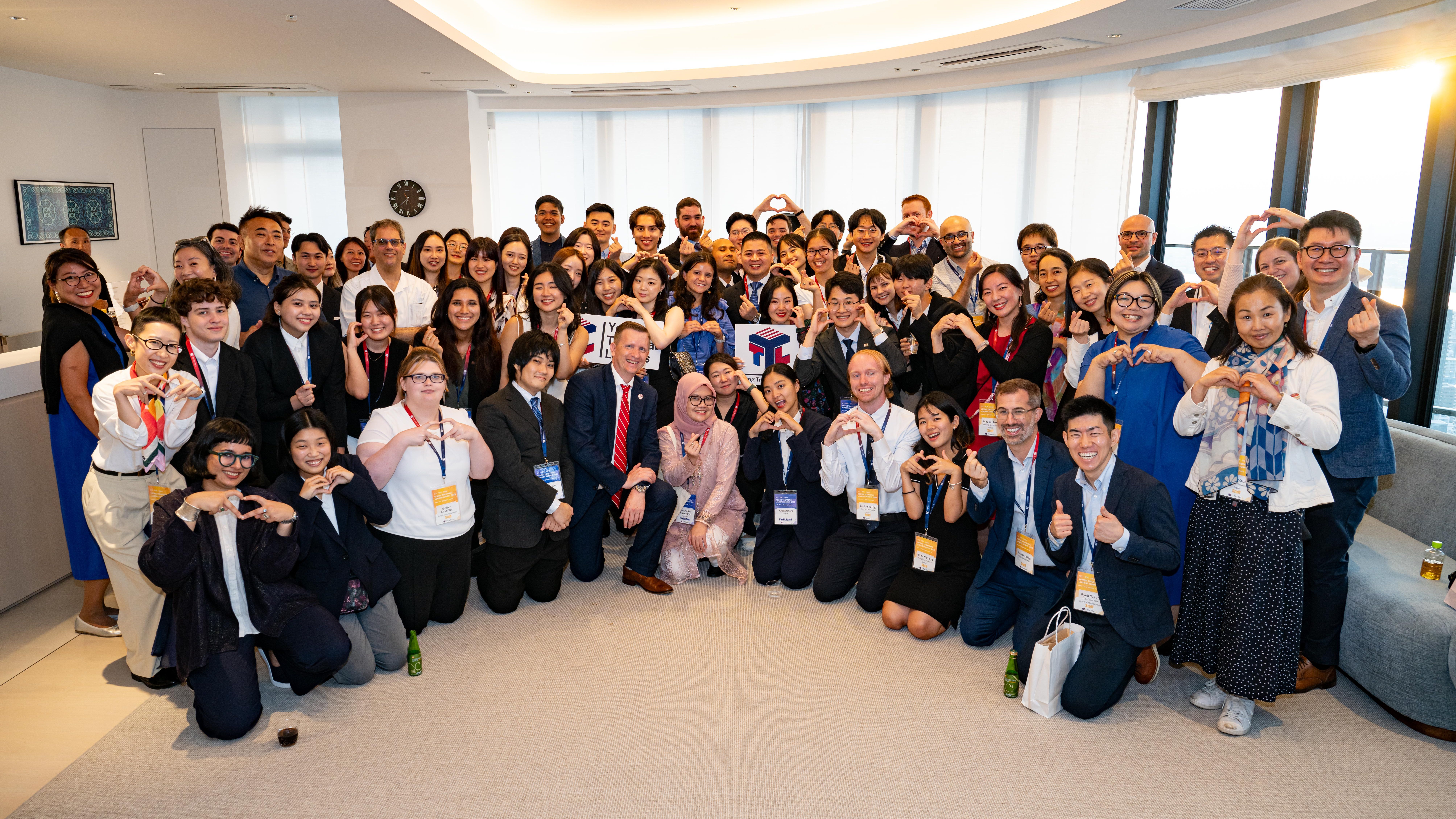 A large group photo of smiling young participants at the U.S.-ROK-Japan Young Trilateral Leaders Summit, showcasing diverse backgrounds and some forming heart shapes with their hands.